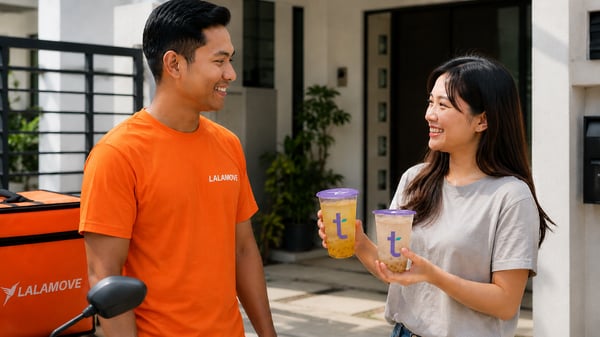 A smiling Malay Lalamove rider in an orange shirt stands beside a Chinese Malaysian woman holding two clear Tealive drinks with purple lids, outside a residential home.