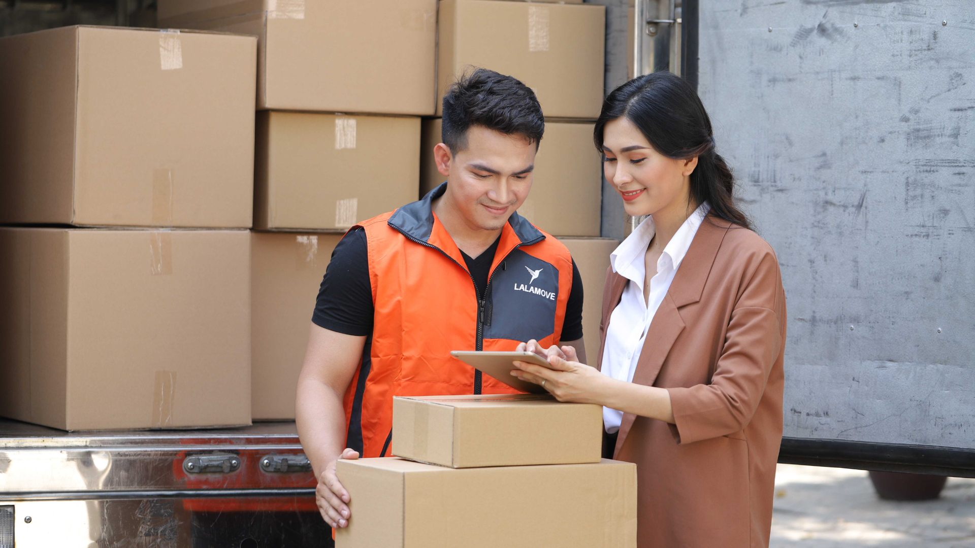 lalamove delivery partner wearing orange shirt is looking at the customers phone while carrying two boxes. behind them are boxes filling a big lorry